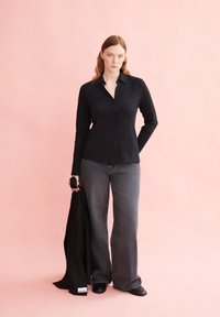 Camicia nera con bottoni e maniche lunghe, abbinata a jeans grigi svasati e una giacca nera. Texture semplici, design minimale, su uno sfondo rosa.