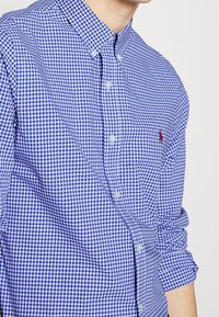 Niebieska i biała koszula w kratę w gingham z kołnierzykiem typu spread i długimi rękawami, z zapięciem na guziki z przodu oraz małym akcentem logo.