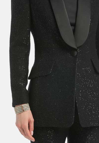 Čierna smokingová bunda s trblietavým textúrovaným materiálom, saténovými klopami, zapínaním na jeden gombík a dvoma prednými vreckami. Obsahuje elegantný náramok.