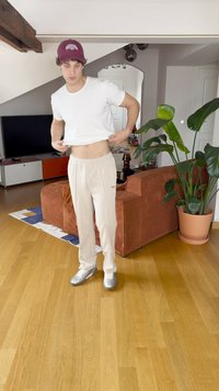 Camiseta branca justa, calças de treino beige e sapatilhas prateadas. O fundo inclui um sofá castanho, uma planta verde e um chão de madeira.