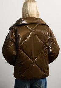 Giacca a vento marrone lucida con un motivo a rombi e un ampio colletto, vista da dietro. Sono visibili dei jeans blu abbinati.
