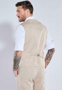 Beige Leinenweste mit slim fit Design, ausgestattet mit einem Rückentrager-Detail und getragen über einem weißen Langarmhemd mit hochgekrempelten Ärmeln.
