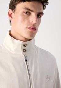 Light beige jacket with a high collar, featuring two button accents and a zipper front. Smooth fabric with a small logo detail on the chest.