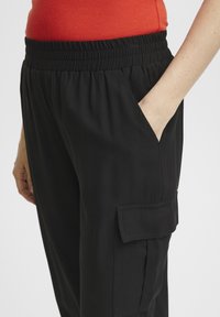 Zwarte broek met een elastische tailleband, zijzakken en een cargozak met flapdetail. Glad stoftextuur, aangesloten bij de taille.