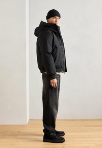 Black puffer jacket with hood, front buttons, elastic cuffs, worn over a cream shirt; paired with dark jeans and black shoes with textured soles.
