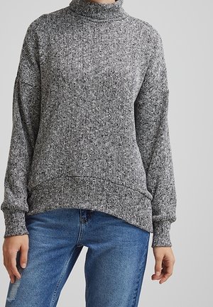 Person wearing a gray textured knit sweater and blue jeans, standing against a plain light gray background.