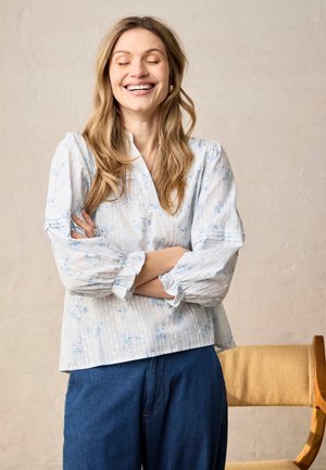 Mujer sonriente con los ojos cerrados, vestida con una blusa blanca con flores y pantalones azules, de pie con los brazos cruzados junto a una silla de madera.