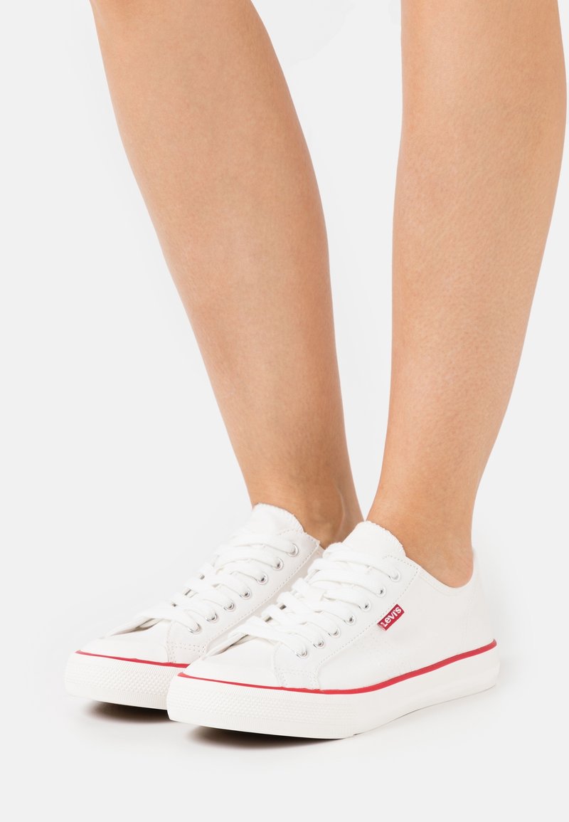 Sneakers bianchi in tela con punta arrotondata, striscia rossa sulla suola e lacci bianchi. La scarpa presenta una tomaia in tessuto testurizzato e un design minimalista.