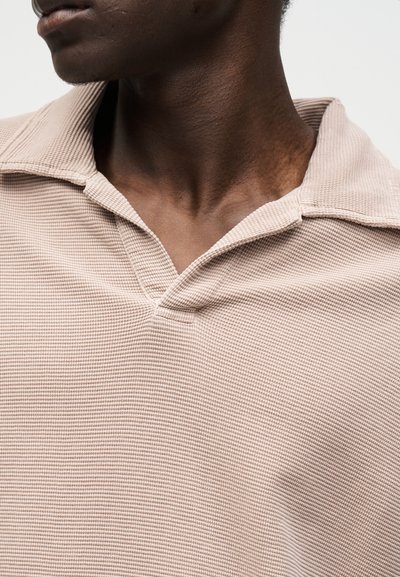 Gros plan d'une personne portant un polo beige texturé avec un col ouvert, montrant le cou et le bas du visage tournés sur le côté.