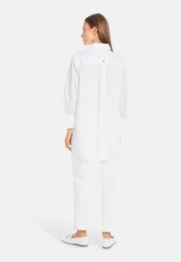 White long-sleeve shirt with a back yoke, featuring buttoned cuff accents. Worn with white pants and casual slip-on shoes.