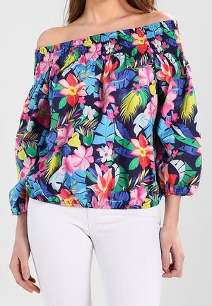 Femme portant un chemisier coloré épaules dénudées avec motifs floraux et feuillages, associé à un pantalon blanc.