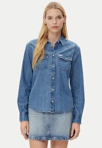 Blonde woman wearing a buttoned blue denim shirt with two chest pockets and a faded blue denim mini skirt, standing against a plain background.