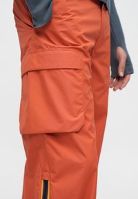 Oranžas kravas bikses no viegla materiāla ar lielu sānu kabatu un vertikālu svītru akcentu potītes rajonā.