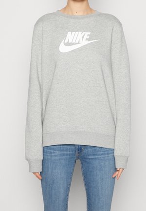 Persoon draagt een lichtgrijze Nike trui met wit logo en blauwe spijkerbroek, staand tegen een effen lichte achtergrond.