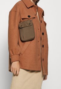 Person iført en brun uldjakke med sorte knapper og en lille olivengrøn crossbody taske over en beige ribbet nederdel.