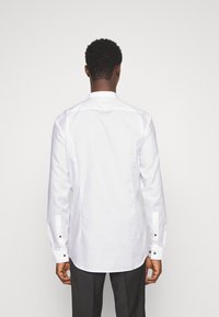 Uomo che indossa una camicia da uomo a maniche lunghe bianca e pantaloni neri, in piedi voltato di spalle di fronte a uno sfondo bianco semplice.