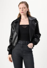 Veste bomber en cuir synthétique noir avec un large col, fermeture éclair, et poignets côtes, associée à un haut noir et un jean foncé.