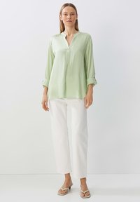 Camicia a maniche lunghe verde chiaro con colletto e pattina dei bottoni, abbinata a pantaloni bianchi a gamba dritta e sandali beige. Tessuto liscio.