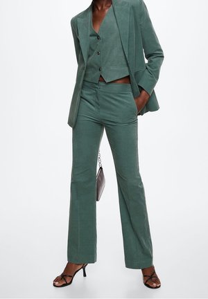 Femme portant un costume vert sur mesure avec un gilet, un pantalon ajusté, des talons noirs à brides, et tenant un petit sac à main sombre.