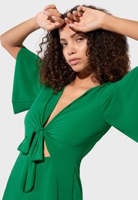 Femme aux cheveux bouclés portant une robe verte à larges manches et nœud à l'avant, posant avec les bras levés au-dessus de sa tête sur un fond clair.