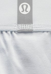 Gaiši pelēks sporta audums ar ribotu struktūru, kurā ir izteikts balts logo un pelēka lenta augšpusē. Redzama elastīga jostasvieta.