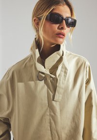 Bēšas krāsas pārāk liela izmēra jaka ar augstu apkakli, ar gredzena detaļu ap kaklu, izgatavota no gludas, vieglas auduma.