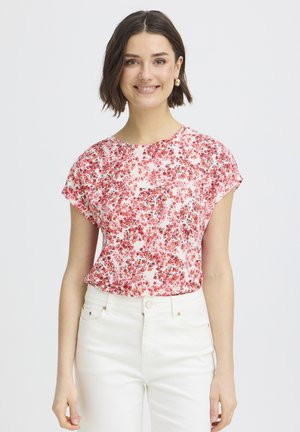 Kobieta o krótkich ciemnych włosach, uśmiechająca się, ubrana w różowo-czerwoną bluzkę w kwiaty z krótkim rękawem oraz białe spodnie z wysokim stanem, na jednolitym tle.