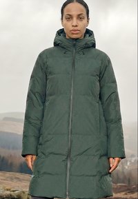 Žena stojaca vonku oblečená v dlhej tmavozelenej prešívanej zimnej bunde s kapucňou, tvárou smerujúcou dopredu s neutrálnym výrazom.