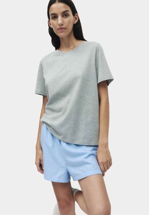 Mujer de cabello largo y oscuro que lleva una camiseta gris lisa de manga corta y pantalones cortos azules claros a cuadros, de pie contra un fondo neutro.