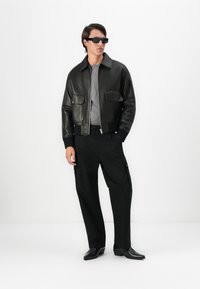 Hombre con chaqueta de cuero negra, camisa gris, pantalones cargo negros, zapatos negros de punta y gafas de sol rectangulares negras, de pie sobre un fondo blanco.
