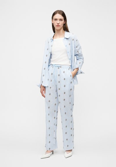 Kvinde står iført lyseblåt pyjamasæt med striber og bi-mønstre, hvid top underneath og hvide spidse hæle på hvid baggrund.