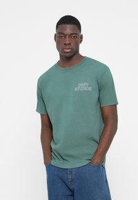 Grøn bomulds t-shirt med korte ærmer. Har "OBEY STUDIOS" logoet i hvid broderi på venstre side. Afslappet pasform, rund halsudskæring.