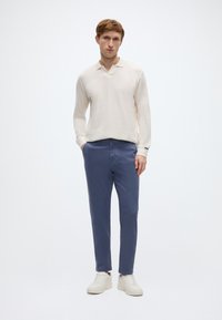 Εκρού μακρυμάνικη μπλούζα με γιακά, συνδυασμένη με στενά παντελόνια σε βαθύ μπλε και λευκά αθλητικά παπούτσια σε ουδέτερο φόντο.