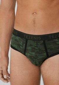 Grüne Camouflage-Unterwäsche mit einem schwarzen Bund, der das "TEZENIS"-Logo trägt. Hergestellt aus weichem Stoff mit glatter Textur und enganliegendem Design.