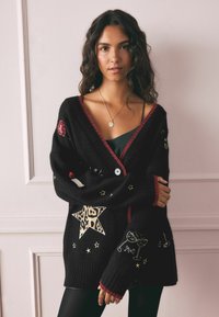 Cárdigan negro con ribetes rojos, que presenta estrellas bordadas, corazones y una estrella con estampado de leopardo. Textura suave y corte oversize.
