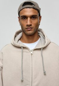 Beige zip-up hoodie met een gevoerde capuchon, grijze trekkoorden en een wit T-shirt eronder. Gdragen met een tan pet en subtiele sieraden.