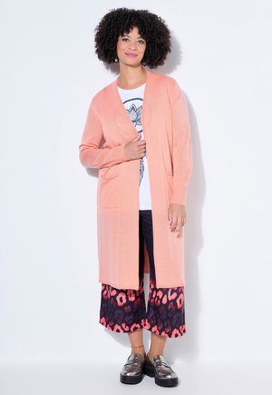 Frau mit lockigem Haar, die eine lange, pfirsichfarbene Strickjacke, ein weißes bedrucktes Hemd, gemusterte weit geschnittene Hosen und metallic Loafers trägt und vor einer weißen Wand steht.