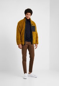 Geel nep-bont jack met een zwart ritssluiting borstzak, gecombineerd met een marineblauwe coltrui en bruine slim-fit broek, en witte sneakers.