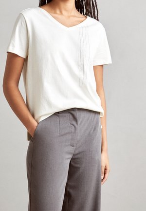 Camiseta de cuello en V en algodón crema claro con mangas cortas, que presenta una línea vertical de pequeños clavos metálicos en el hombro izquierdo. Combinada con pantalones gris ajustados.