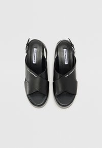 Sandali slingback neri in pelle con design a tracolla incrociata, suola piatta e texture liscia. Presentano il marchio sulla soletta. Chiusura con fibbia regolabile.