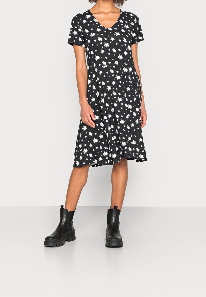 Femme portant une robe noire au genou avec un imprimé floral blanc et des bottines noires épaisses, debout devant un fond uni.