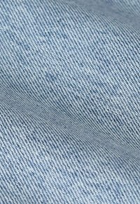 Un tissu en denim bleu clair présente une surface lisse et texturée avec de fins motifs en chevrons et des variations subtiles de couleur.