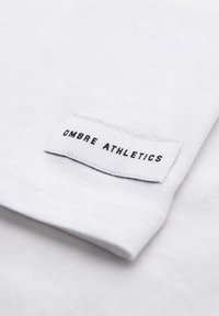 Tissu blanc avec une étiquette blanche rectangulaire cousue, affichant le texte noir "OMBRE ATHLETICS" en lettres majuscules.