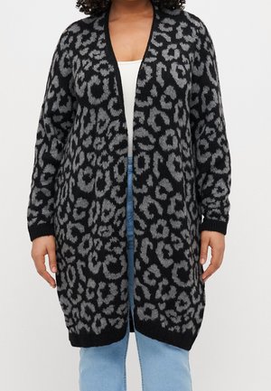Femme portant un long cardigan noir et gris à imprimé léopard ouvert sur le devant, par-dessus un haut blanc et un jean bleu clair.