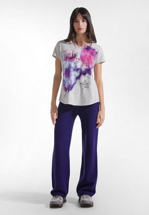 Vrouw met lang donker haar, die een wit T-shirt draagt met roze en paarse spatten, donkerblauwe broek en grijze sportschoenen, staand op een witte achtergrond.