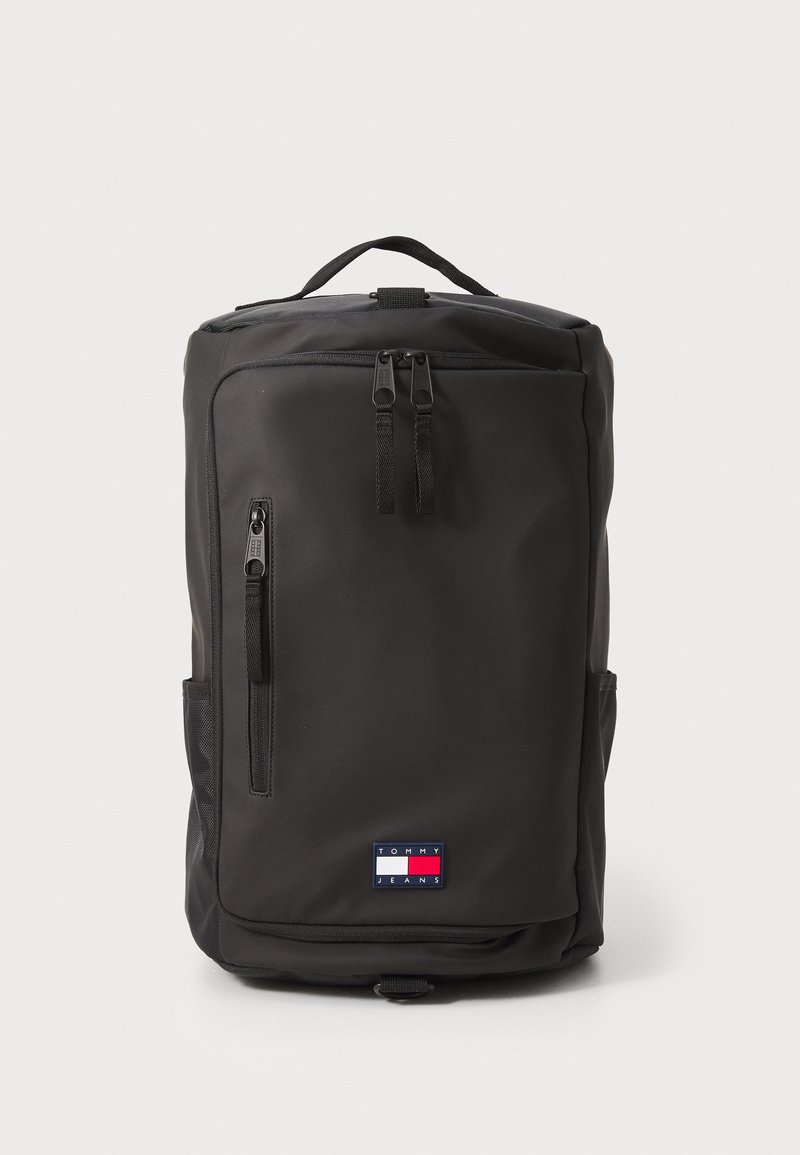 Black backpack with two top zippers, side pockets, and a small logo patch in red, white, and blue. Smooth texture and rectangular shape.