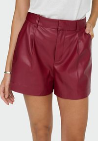 Bordeauxfarbene Kunstleder-Shorts mit hoher Taille, gefaltetem Vorderteil, seitlichen Taschen und einer glatten, glänzenden Textur. Maßgeschneiderte Passform mit klaren Linien.