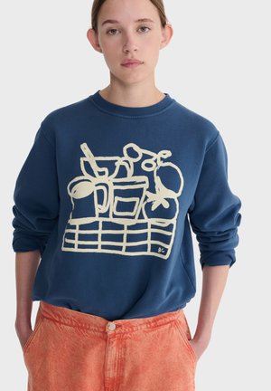 Young person wearing a blue sweatshirt with white abstract kitchenware print and orange pants, standing with hands in pockets.