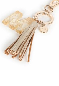 Portachiavi con una lettera "M" dorata e texturizzata, un pon-pon beige e un piccolo tag rotondo inciso con "MIRIADE", attaccato con un anello color oro rosa.