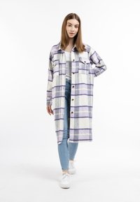 Giacca camicia a quadri bianca, grigia e viola con bottoni, due tasche anteriori e un taglio lungo e dritto. Indossata sopra un top bianco e jeans.